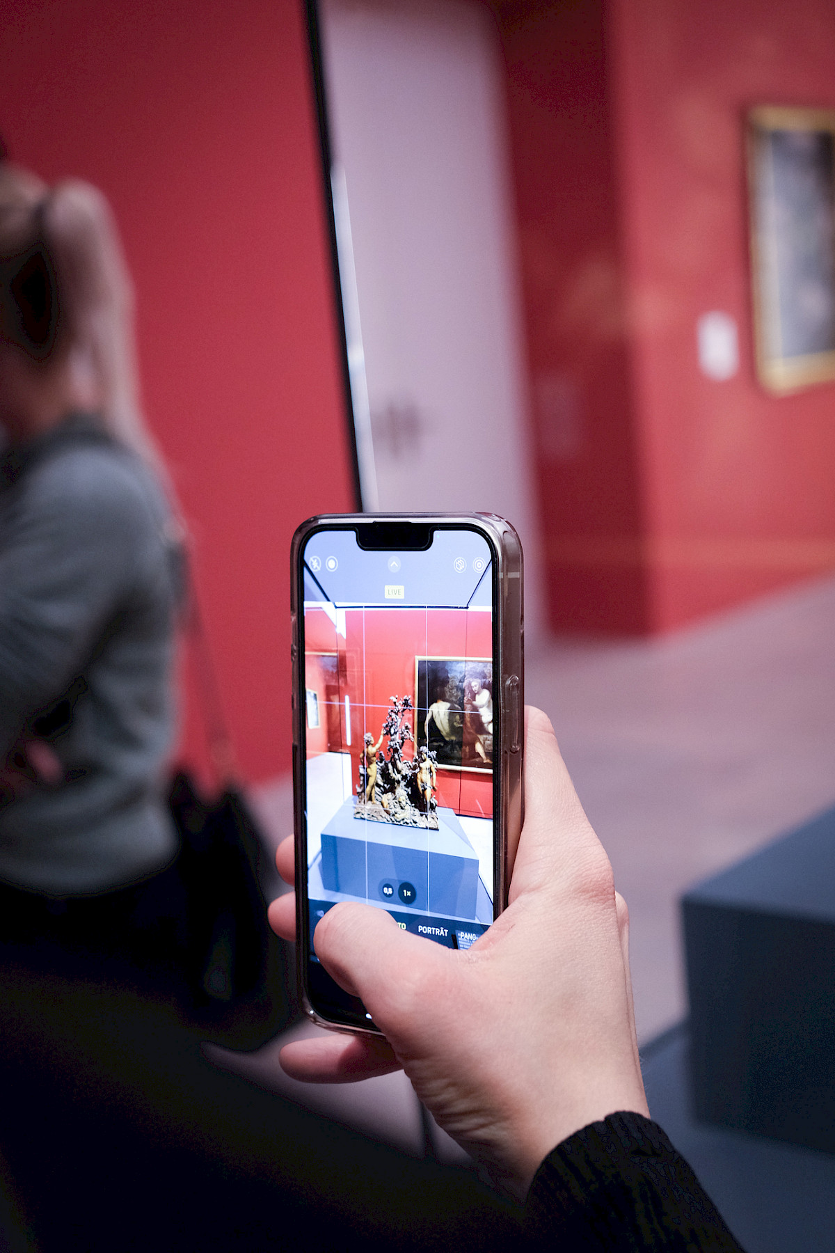 Hand hält Smartphone, das eine Skulptur in einem Museum vor roter Wand fotografiert.